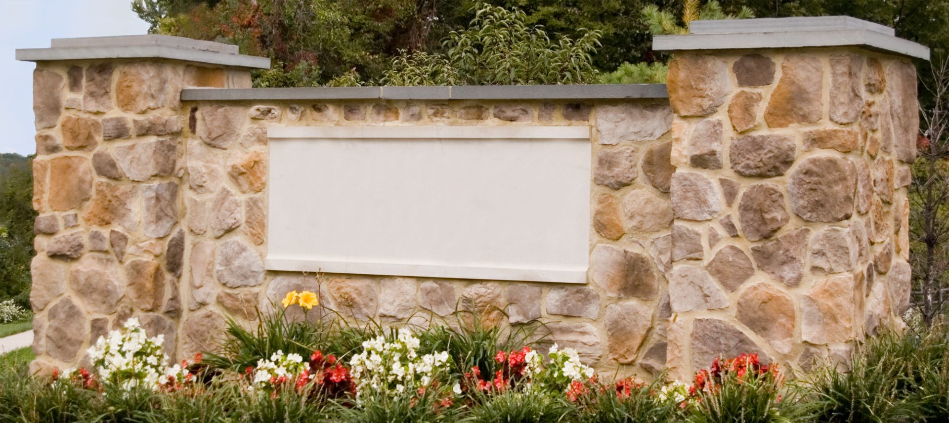 A sign in front of a stone wall with flowers around it.